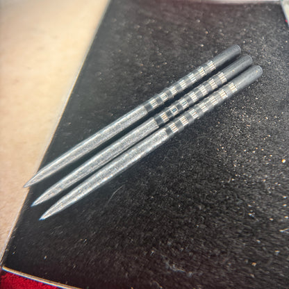 Three silver metallic dart shafts with black and white striped bands near the tips, resting on a textured black surface.