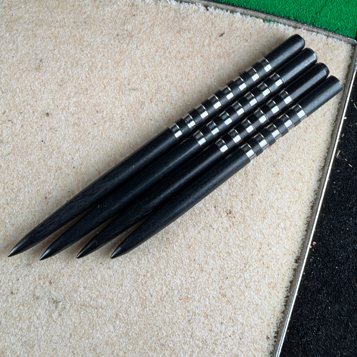 Four black darts with silver bands arranged diagonally on a textured beige surface.